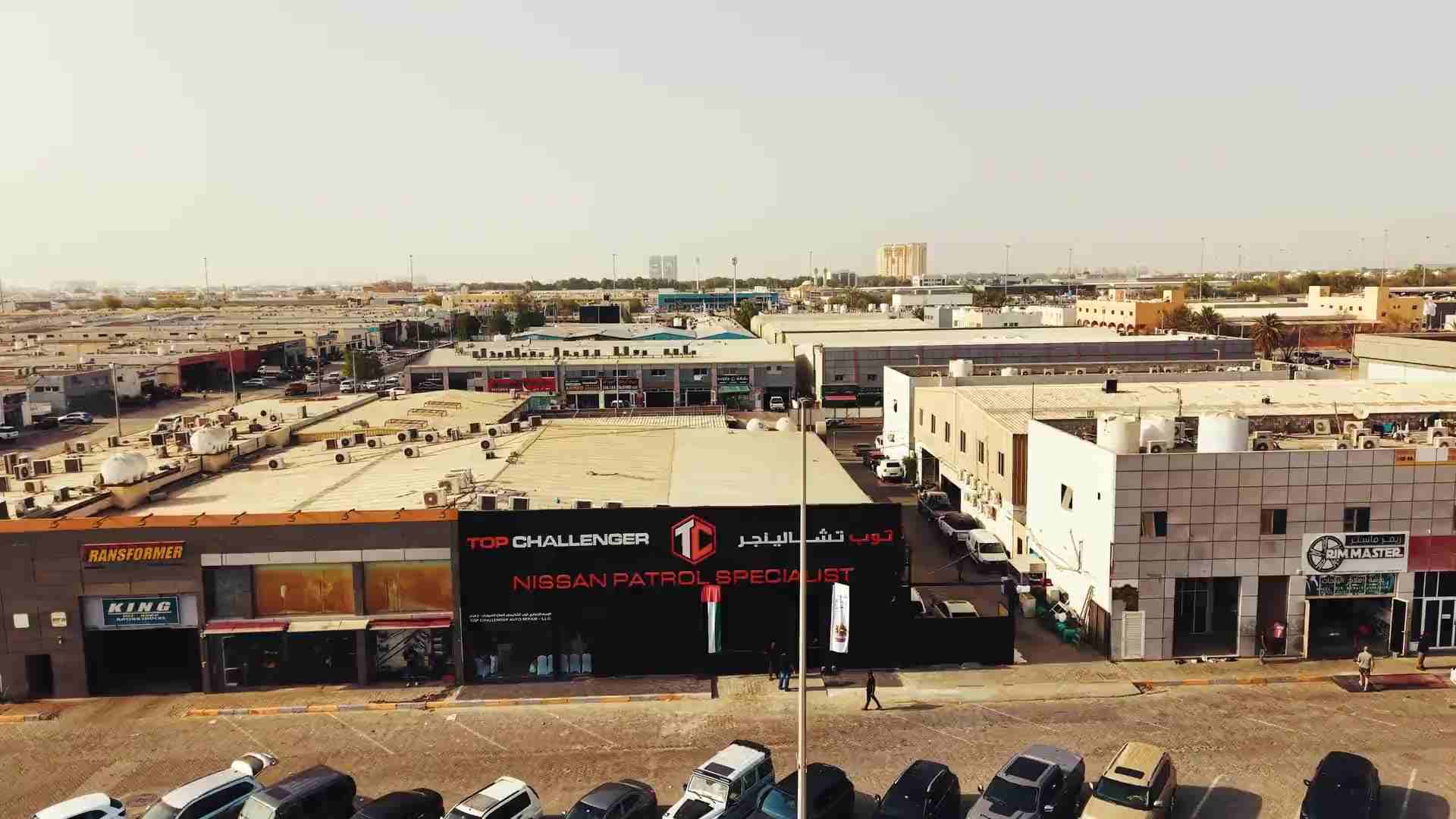 The Top Challenger workshop in Mussafah Industrial, Abu Dhabi, seen from the air: a black frontage carrying the name in English and Arabic either side of the orange TC hexagon, with Nissan Patrol Specialist beneath it in red, among the low workshop units and flat roofs of the industrial area